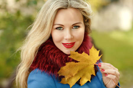 Luxury blonde girl with beautiful hair in a coat holding a maple leaf in autumnの写真素材