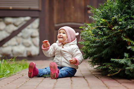 Very charming beautiful little girl with big brown eyes sits and smiles on the background of wooden houses and trees in autumnの写真素材