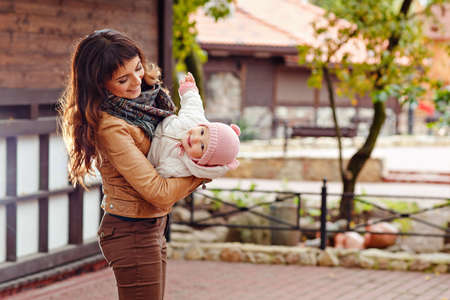 A beautiful brunette mom is holding his head down a charming beautiful little girl smiling on background of house in autumnの写真素材