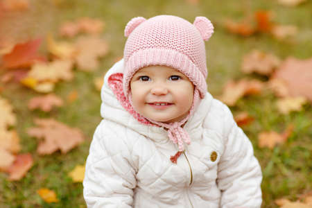 Very charming beautiful little girl with big brown eyes and pink hat sitting on grass and smiling on background of yellow maple leaves in autumn, close-upの写真素材