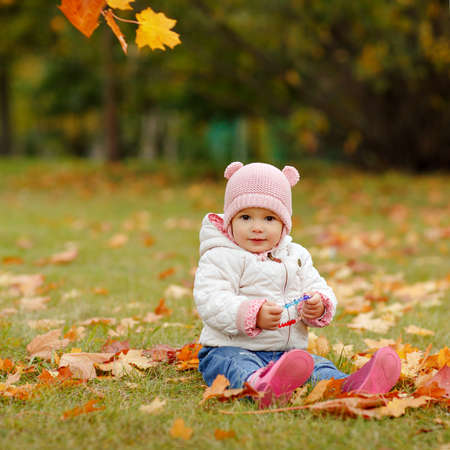 Very charming beautiful little girl with big brown eyes and pink hat sitting on grass and smiling on background of yellow maple leaves in autumnの写真素材