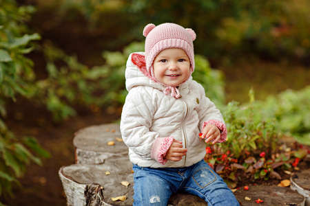 Very charming beautiful little girl with big brown eyes sits and smiles on the background of green leaves in autumnの写真素材
