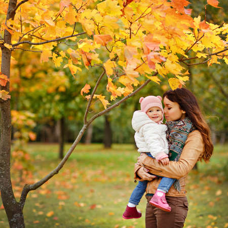 Very charming beautiful brunette mom in a brown jacket holding a little girl her daughter on the background of yellow leaves of maple in autumnの写真素材