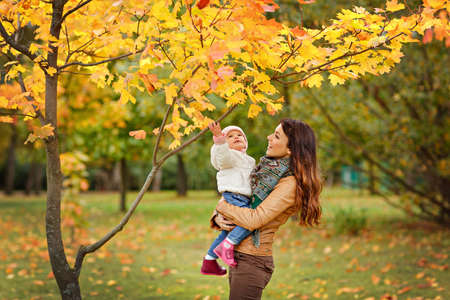 Very charming beautiful brunette mom in a brown jacket holding a little girl her daughter and smiles happily on the background of yellow leaves of maple in autumnの写真素材