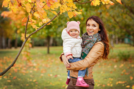Very charming beautiful brunette mom in a brown jacket holding a little girl her daughter and smiles happily on the background of yellow leaves of maple in autumnの写真素材