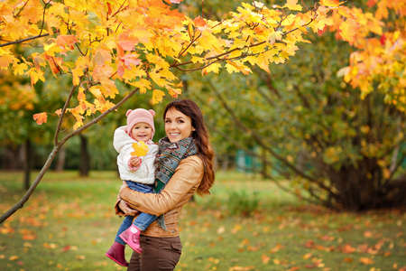 Very charming beautiful brunette mom in a brown jacket holding a little girl her daughter and smiles happily on the background of yellow leaves of maple in autumnの写真素材