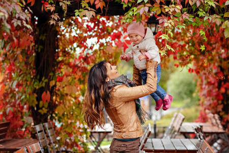 Very charming beautiful brunette mom in a brown jacket throws up little girl daughter on background of red leaves of grapes in autumnの写真素材