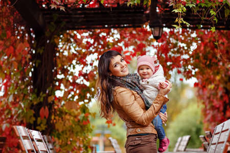 Very charming beautiful brunette mom in a brown jacket holding a little girl her daughter and smiles happily on the background of red leaves of grapes in autumnの写真素材