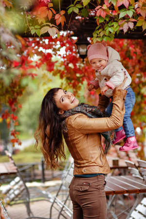 Very charming beautiful brunette mom in a brown jacket throws up the little girl her daughter and smiles happily on the background of red leaves of grapes in autumnの写真素材