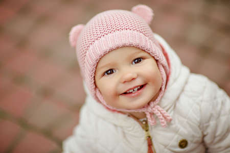 Very charming beautiful little girl with big brown eyes in a pink cap smiles in autumn, close-upの写真素材