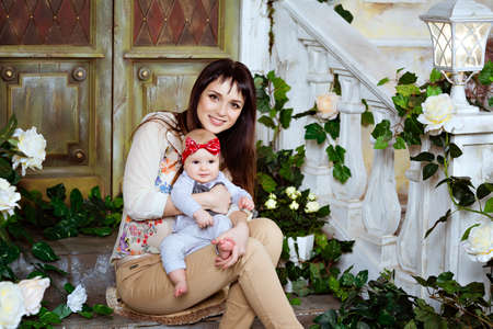 Happy and beautiful mom brunette is holding his adorable baby daughter, sitting on the stairs of an old houseの写真素材
