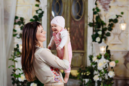 Happy and beautiful mom brunette holding a charming little daughter baby and laughing together, in the interiors of the house in beigeの写真素材