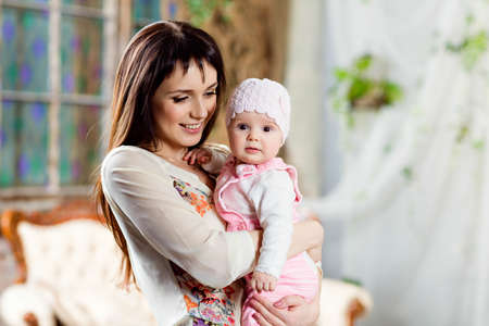 Happy and beautiful mom brunette holding a charming little daughter baby in the interior in beige tonesの写真素材