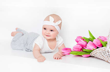 Small very cute wide-eyed smiling baby girl lying on her tummy on a white background in the Studio, next to the tulipsの写真素材