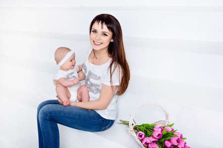 Beautiful and happy mom brunette in white t-shirt and jeans holding a little smiling baby daughter on a white background with tulipsの写真素材