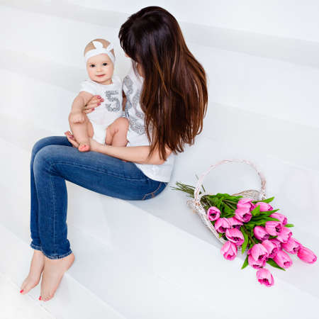 Beautiful and happy mom brunette in white t-shirt and jeans holding on her knees a little smiling baby daughter on a white background with tulipsの写真素材