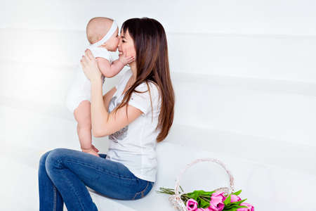 little smiling baby daughter bites his nose mom brunette in white t-shirt and jeans on a white background in the Studio, next to the tulipsの写真素材