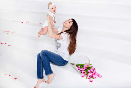 Beautiful and happy mom brunette in white t-shirt and jeans throws up a little smiling baby daughter on a white background with tulipsの写真素材