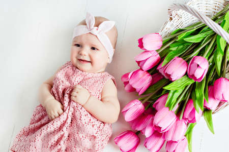 Small very cute, big-eyed little baby girl in a pink dress lying on a white background in the Studio about pink tulipsの写真素材