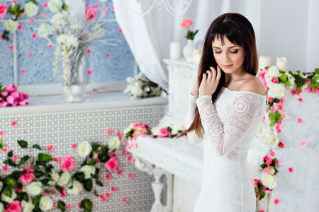 very beautiful sensual brunette girl with long straight hair is standing with closed eyes in a white lace dress on the background of spring flower decorations and a white pianoの写真素材