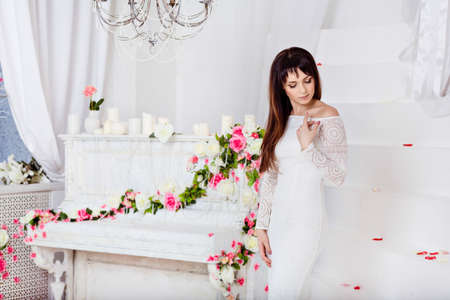 very beautiful sensual brunette girl with long straight hair is in a white lace dress on the background of spring flower decorations and a white pianoの写真素材