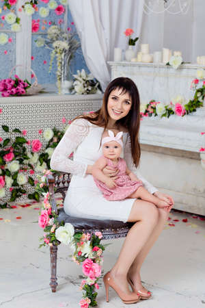 Beautiful and happy mom brunette in a white dress holding on the handles a little girl daughter on the background of the spring interior with flowers in full growthの写真素材