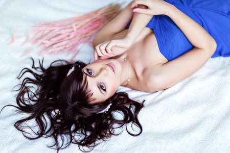 Portrait of a cute sensual beautiful young girl brunette in a blue dress, lying on a white bedの写真素材