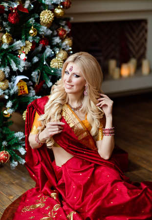 Very beautiful blonde girl in Indian red saree sitting on the floor around the Christmas treeの写真素材