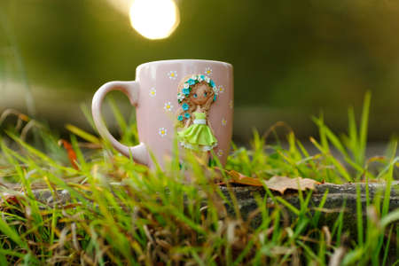 Pink mug with decorations in the form of a girl in a dress made of polymer clay on nature backgroundの写真素材
