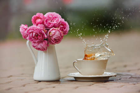 Splashes of tea from a white mug, next to a vase of pink roses in the summer