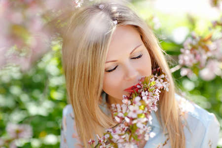 Portrait of a beautiful young blonde woman with long hair in a blue dress on a background of pink cherry blossoms in spring, close-up with eyes closedの写真素材