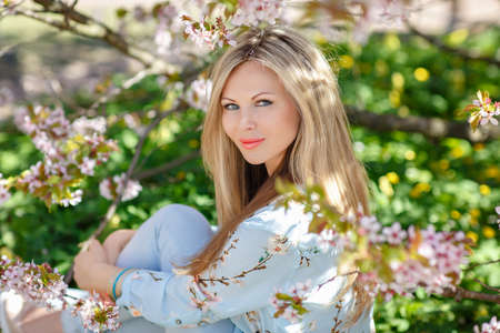 Portrait of a beautiful young blonde woman with long hair in a blue dress on a background of pink cherry blossoms in springの写真素材