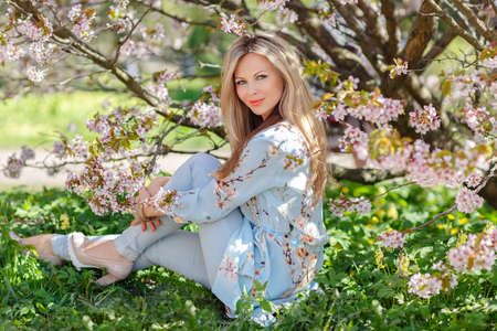 Portrait of a beautiful young blonde woman with long hair in a blue dress on a background of pink cherry blossoms in springの写真素材