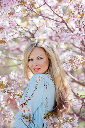 Portrait of a beautiful young blonde woman with long hair in a blue dress on a background of pink cherry blossoms in springの写真素材