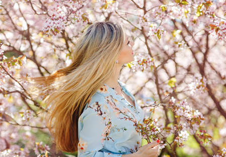 Beautiful blonde girl with long hair in a blue dress on a background of pink cherry blossoms in springの写真素材