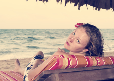 Beautiful woman sitting by the ocean with a flower in her hair, looking at the cameraの写真素材