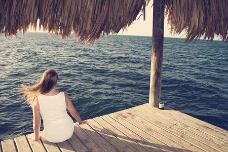Woman in white dress sits on a dock and looks out at the oceanの写真素材