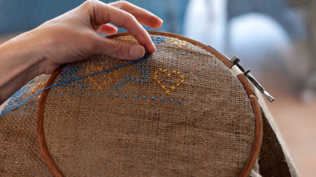 Female hand hold needle with blue thread and embroider cross stitch pattern on rough burlap fabric...