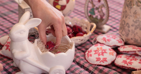 A hand is gently placing bright red Easter eggs inside a ceramic bunny vessel. Quail eggs are arranged nearby, creating a cheerful atmosphere for the holiday celebration.の写真素材