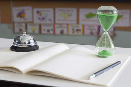 Hourglass with green sand and front desk bell on white notebook with pencil, selective focus on...