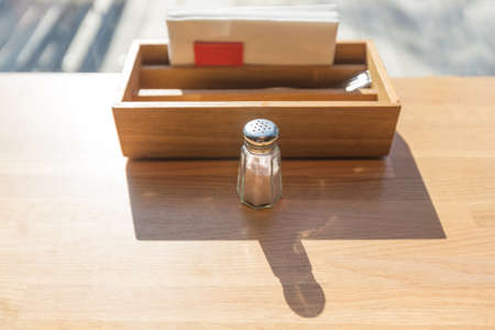 Glass salt shaker on a wooden table in a restaurant placed next to a wooden box of folks and napkins, backlit by morning sunlight casting long shadow, selective focus on the shaker's lid with shallow depth of fieldの写真素材
