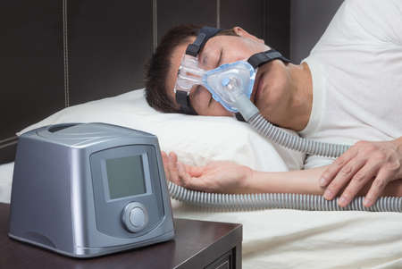 Asian man with sleep apnea using CPAP machine, wearing headgear mask connecting to air tubeの写真素材