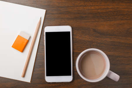 Blank white notebook, pencil, eraser, phone, and hot drink on wooden deskの写真素材