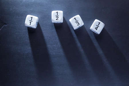 white fraction math number dices on blackboard, good for background and teaching blogs, with harsh lighting showing long dices' shadows, and selective focus on dices, room for copyspace on top of frameの写真素材