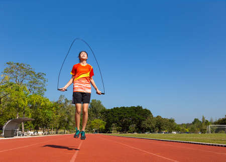 young female asian athlete rope jumping outdoor on running track at football, or soccer, field with trees and clear blue sky in backgroundの写真素材
