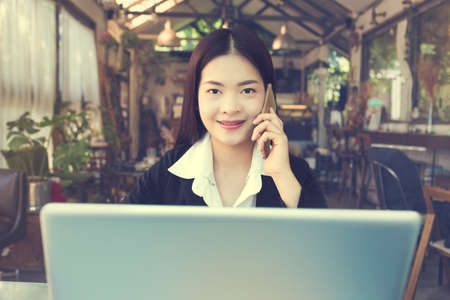 Smiling young asian executive business woman working on her computer and talking on her phone indoor, golden hour lighting coming through the large windows, vintage retro style lookの写真素材