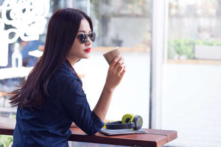 attractive young woan wearing casual clothes and sunglasses, holding cup of coffee, waiting for her friend at a coffee shopの写真素材