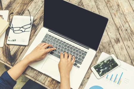 Top view of female hands typing on laptop keyboard with smart phone, eyeglasses, and paper notebooks on wooden table, in vintage color styleの写真素材