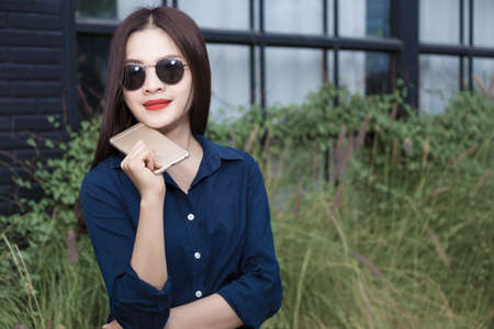 young asian woman in casule clothes wearing sunglasses holding cellphone waiting outside a coffee shop, blue color tone, room for copy spaceの写真素材