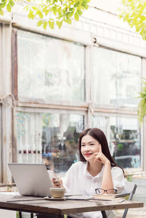 happy young asian woman thinking and enjoying her coffee while using her computer and smart phoneの写真素材
